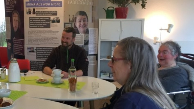 Auf dem Bild sitzen zwei Frauen und ein Mann an einem runden Tisch. Im Hintergrund steht die Wanderausstellung der KSL NRW. Auf dem Tisch stehen Getränke und Kekse.