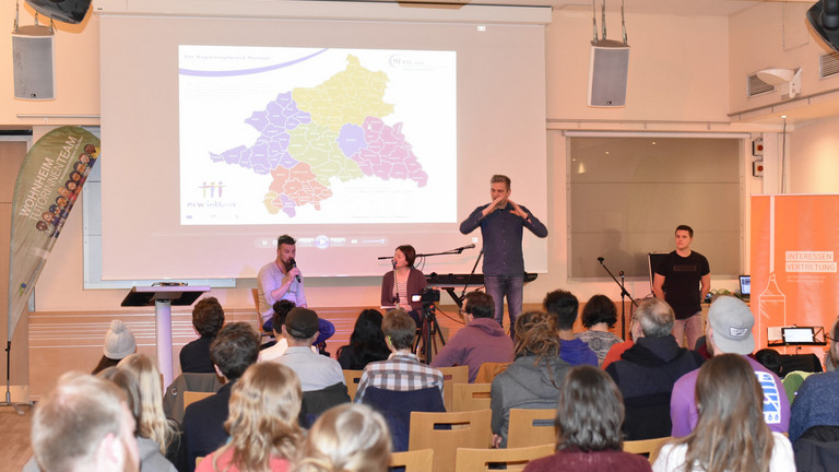 Auf dem Bild sind die Hinterköpfe des sitzenden Publikum zu sehen. Auf der davor liegenden Bühne sitzen links ein Mann und rechts eine Frau auf Stühlen. Rechts neben der Frau steht ein Gebärdensprachdolmetscher. Er hat eine Jeans und ein blaues Hemd mit Muster an. Im Hintergrund zu der Bühne hängt eine Leinwand auf dieser ist die Landkarte des Regierungsbezirks Münster in bunten Farben zu sehen. Das Licht im Raum ist gedämmt.