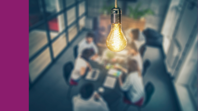 Foto von mehreren Menschen, die an einem Tisch sitzen im Hintergund. Eine Glühlampe ist im Vordergrund des Bildes.