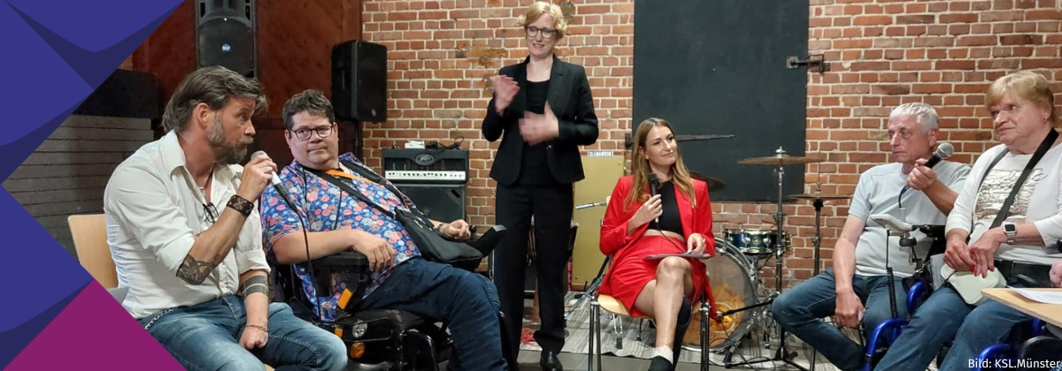Oliver Schneider spricht in einer Gesprächsrunde mit zwei Menschen, die einen Rollstuhl nutzen, einer Moderatorin, einer Assistenzkraft und einer DGS-Dolmetscherin