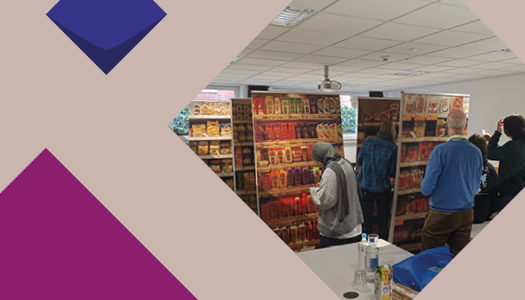 Eine lila Kachel und ein Foto von einem Seminarraum. Es sind mehrere Stellwände neben- und hintereinander aufgestellt, mit denen Supermarktregale nachgeahmt werden. Auf einem sind verschiedene Shampooflaschen abgebildet, auf der anderen Wurstverpackungen. Fünf Personen stehen verteilt vor den Stellwänden und begutachten sie.