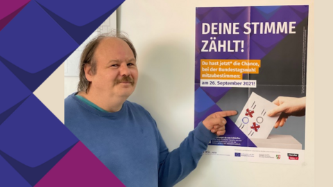 Foto von Gerd Ziegler. Er zeigt auf das Wahl-Plakat der KSL NRW