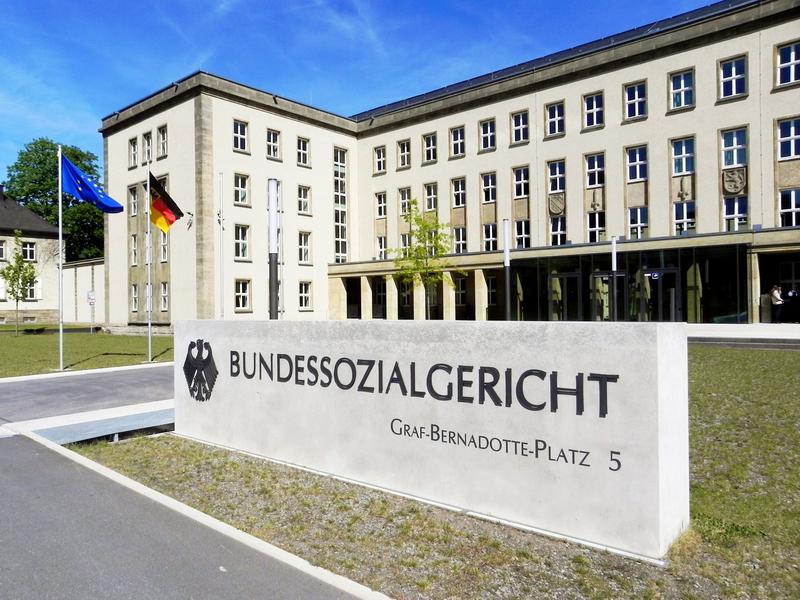 Das Foto zeigt den Eingangsbereich zum Bundessozialgericht. Eingrußes weißes Gebäude. Davor befindet sich eine weiße Tafel mit der Aufschrift Bundessozialgericht.