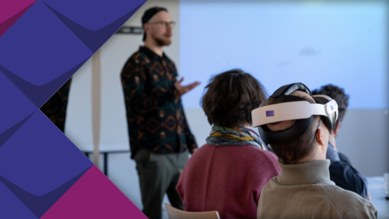 Foto einer Workshop-Situation. Eine sitzende Person im Fokus trägt eine VR-Brille und neigt den Kopf leicht nach oben. Im Hintergrund sitzen zwei weitere zuhörende Personen. Marcel Scharf vom KSL.Münster ist als sprechender Referent unscharf im Hintergrund zu erkennen.